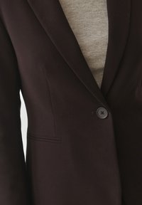 Dark brown blazer with a smooth texture, featuring a single button closure and a tailored fit. Includes a subtle side pocket detail.
