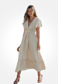 Robe maxi beige avec un décolleté en V profond, des manches courtes bouffantes et un détail de taille en dentelle crochetée, présentant une silhouette fluide et un ourlet à volants.