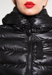 Czarna kurtka puchowa o błyszczącym wykończeniu, z zamkiem błyskawicznym i kapturem ze sznurkiem. Posiada akcenty w postaci guzików oraz lekki, pikowany materiał.