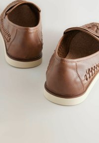 Zapatos de cuero marrón tipo mocasín con suelas blancas, que presentan acentos laterales texturizados y un acabado suave. El diseño incluye una punta redondeada y un perfil bajo.