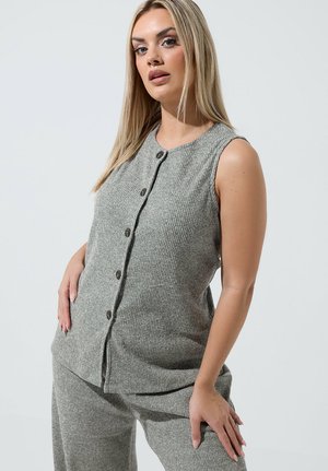 Mujer rubia con top gris sin mangas de canalé con botones y pantalones a juego, posando con la mano en la cadera contra un fondo claro y liso.