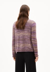 Personne avec de longs cheveux bruns, portant un pull rayé violet et marron et un pantalon noir, se tenant de dos face au spectateur.