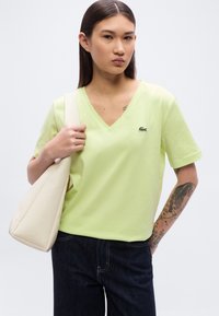T-shirt vert citron à manches courtes et col en V, en coton, avec un petit logo brodé sur la poitrine. Porté avec un jean denim foncé et un sac beige.