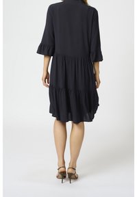 Dunkelblaues Kleid mit dreiviertellangen Rüscheärmeln, gestuftem Design und lockerer Passform, kombiniert mit sandalenartigen hohen Schuhen mit Riemchen.