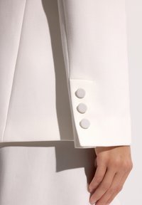 Rękaw białej marynarki z trzema guzikami obłożonymi tkaniną, spoczywający blisko dłoni na jasnym tle.