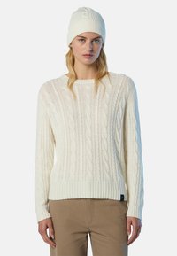 Donna bionda che indossa un maglione bianco a trecce, un berretto di maglia bianco abbinato e pantaloni beige, in piedi contro uno sfondo semplice.
