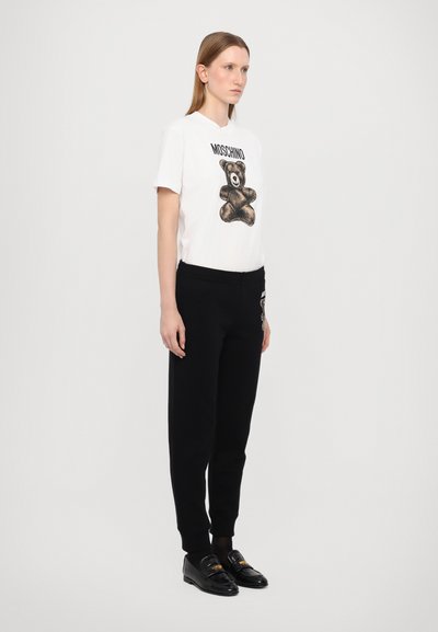 Witte T-shirt met een zwart teddybeer graphic en de tekst "Moschino", gecombineerd met zwarte sweatpants en zwarte instappers met gouden accenten.