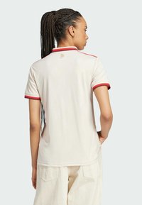 T-shirt polo crème avec des accents rouges, manches courtes et une finition texturée. Présente un petit logo à l'arrière. Porté avec un pantalon de couleur claire.