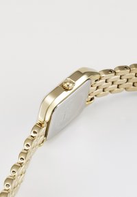 Montre-bracelet en or avec un visage rectangulaire, une chaîne en or liée et une couronne proéminente sur le côté. Surface lisse et finition métallique.