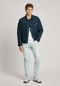 Chaqueta acolchada azul marino con diseño de cuello, botones a presión, dos bolsillos en el pecho, combinada con jeans azul claro y zapatillas blancas.
