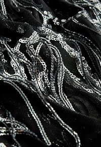 Czarna tkanina ozdobiona srebrnymi cekinami, tworząc teksturowany, błyszczący wzór. Cekiny tworzą falujące, faliste kształty na materiale.