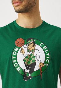 Πράσινο βαμβακερό T-shirt με λογότυπο των Boston Celtics που απεικονίζει έναν λευκό μουσακά, μπάλα μπάσκετ και μοτίβο τριφύλλι. Απλή στρογγυλή λαιμόκοψη.