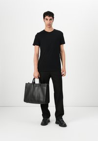 Geantă tote din piele neagră, cu două mânere superioare, textură netedă și un design structurat; purtată cu un tricou negru și pantaloni tailoring.