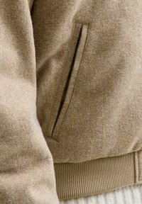 Chaqueta de mezcla de lana marrón con textura suave, que presenta un bolsillo lateral y un dobladillo acanalado. El diseño es casual y ajustado.