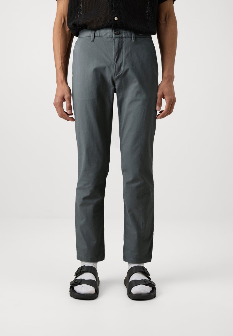 Homme portant un pantalon gris droit, des chaussettes blanches, des sandales noires à boucle et une chemise noire à manches courtes sur un fond uni.