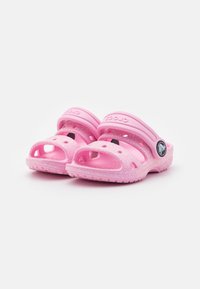 Vaaleanpunaiset Crocs-sandaalit kiiltävällä pinnalla, joissa on useita ilmanvaihtoaukkoja ja muotoiltu pohja. Kestävä hihna kengän päällä.