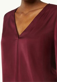 Blusa de seda burdeos con un profundo escote en V y textura suave, que presenta un ligero plisado cerca del cuello y un corte relajado.