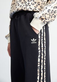 Pantaloni della tuta neri con strisce laterali a stampa leopardata, vita elasticizzata e tasche. Presenta un logo Adidas sull'anca sinistra.