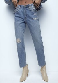 Jeans de mezclilla azul claro con parches desgastados, corte recto, cintura alta y dobladillos deshilachados, combinados con botines beige de punta.