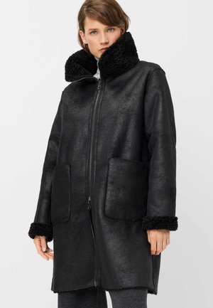 Woman wearing a long black shearling coat with large collar and cuffs, standing against a plain light background.