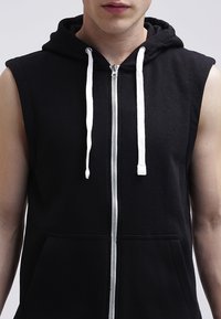 Sudadera sin mangas negra con cremallera frontal, cordones blancos y bolsillos tipo canguro. Textura de tela suave, diseño casual, estilo minimalista en general.