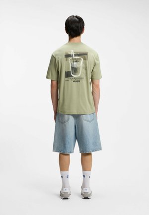 Uomo di spalle che indossa una maglietta verde chiaro con una grafica di bevanda e pantaloncini di jeans, calzini bianchi e scarpe da ginnastica su uno sfondo bianco semplice.