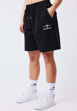 Persona che indossa pantaloncini neri con il logo PXP, calzini bianchi fino a metà polpaccio con logo nero e scarpe da ginnastica bianche, in piedi con una mano in tasca.