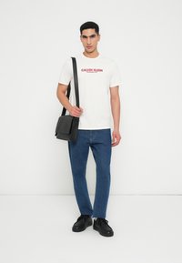 Νεαρός άνδρας που φοράει άσπρη μπλούζα Calvin Klein, μπλε τζιν, μαύρα παπούτσια, κρατώντας μαύρη τσάντα χιαστί με λουρί στον ώμο, μπροστά σε λευκό φόντο.