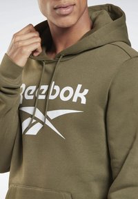 Olivgrüner Reebok-Hoodie aus weichem Material mit einer frontalen Tasche, ausgestattet mit einem großen weißen Logo und einer Kordelzugkapuze.