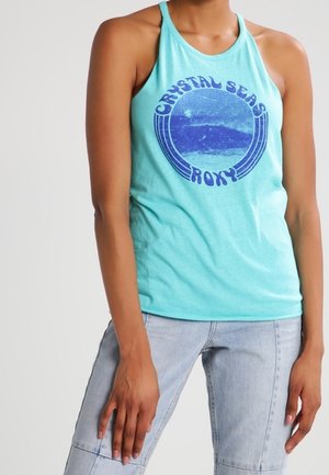 Kvinde iført en turkis ærmeløs tanktop med en blå cirkulær "Crystal Seas Roxy" grafik og lyseblå jeans.