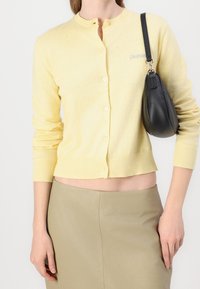 Yellow cropped cardigan with a round neckline, button front, ribbed cuffs, and hem. Paired with a smooth olive-green skirt and a black bag.