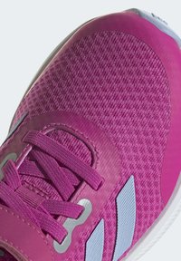 Zapatilla deportiva rosa con un upper texturizado, cordones planos y detalles plateados, con una suela de goma lisa y un diseño de puntera redondeada.