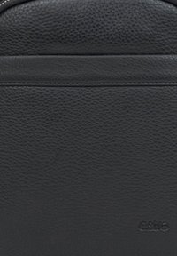 Černá kožená taška s texturou jemného písku, hladkými akcenty a raženým logem v dolním pravém rohu. Minimalistický design bez kovových prvků.