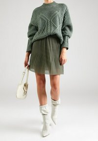 Vihreä neulottu neule tekstuurisilla yksityiskohdilla, laskostettu oliivinvihreä hame, valkoiset löysät nilkkurit ja kermahtava käsilaukku.