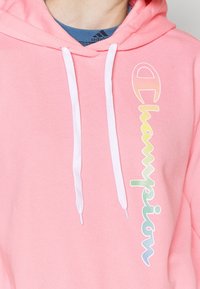 Rosa bomullshuvtröja med vita dragkedjor. "Champion"-logotypen i en gradientdesign löper vertikalt längs framsidan. Mjuk textur.