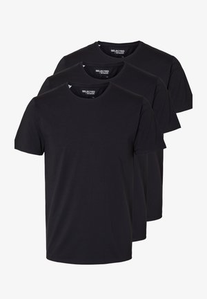 Junger Mann trägt ein schwarzes T-Shirt und schwarze Jeans, hält eine schwarze Tragetasche in einer Hand und steht vor einem einfarbigen hellen Hintergrund.