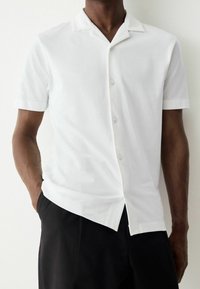 Weißes kurzärmeliges Hemd mit Knöpfen, spitzem Kragen, glatter Textur und leicht geschwungenem Saum, kombiniert mit schwarzen Hosen.