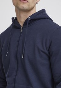 Navyblauwe ritshoodie van zachte stof, met een capuchon, trekkoorden en hardware in zilverkleur. Glad textuur met een ontspannen pasvorm.