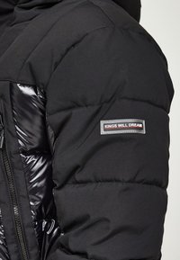 Svart pufferjacka med en glansig, quiltad textur. Har dragkedja och en axelpatch märkt "KINGS WILL DREAM" i grått och rött.