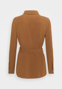 Giacca marrone aderente con colletto, vita con cintura e maniche lunghe. Realizzata in materiale testurizzato, presenta cuciture verticali e dettagli con bottoni.