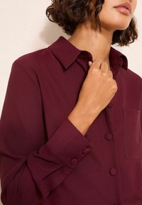 Bordowa bluzka z kołnierzykiem, zapinana z przodu na guziki, z długimi rękawami. Posiada kieszeń oraz gładki, teksturowany materiał.