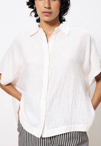 Weißes Button-up-Hemd mit lockerer Passform, aus strukturiertem Stoff gefertigt. Mit kurzen Ärmeln und einem klassischen Kragen, kombiniert mit gestreiften Hosen.