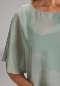 Top in chiffon verde chiaro con maniche corte, caratterizzato da una superficie liscia e sottili motivi floreali in toni più chiari lungo il tessuto.