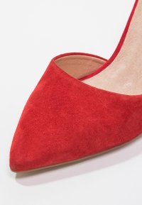 Chaussure à talon haut en suède rouge avec un bout pointu et des côtés ouverts, dotée d'une doublure intérieure lisse et d'un talon fin.