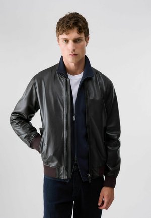 Giovane uomo con capelli ricci che indossa una giacca di pelle nera sopra una felpa blu navy con zip e una camicia bianca, in piedi davanti a uno sfondo grigio uniforme.