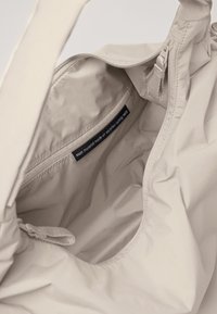 Sac fourre-tout beige avec un extérieur en nylon recyclé lisse. L'intérieur comprend une poche zippée et un tissu étiqueté indiquant l'origine du matériau.