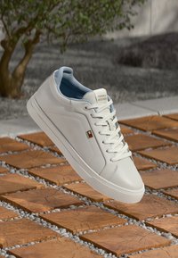 Sneaker bianco Tommy Hilfiger con lacci, interno blu e logo di lato, posizionata sopra piastrelle di legno all'aperto.