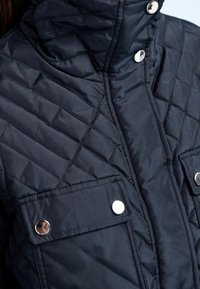 Genähte marineblaue Jacke mit hohem Kragen, ausgestattet mit silbernen Druckknöpfen und zwei Brusttaschen. Glattes, leichtes Material mit dezentem Glanz.