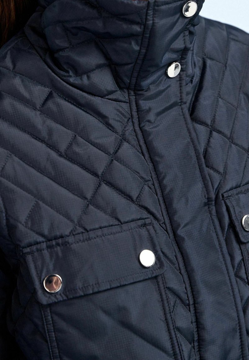 Genähte marineblaue Jacke mit hohem Kragen, ausgestattet mit silbernen Druckknöpfen und zwei Brusttaschen. Glattes, leichtes Material mit dezentem Glanz.