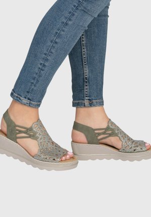Feet wearing open-toe olive green wedge sandals with cutout and rhinestone design, paired with blue skinny jeans.